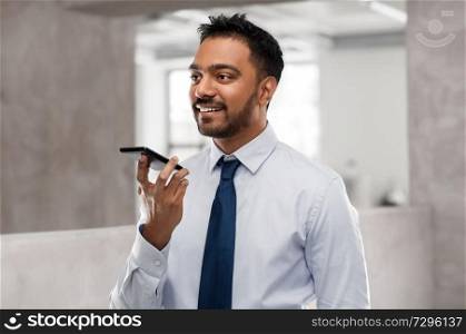 business, communication and technology concept - smiling indian businessman using voice command recorder on smartphone over office background. businessman using voice command on smartphone