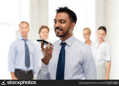 business, communication and technology concept - smiling indian businessman using voice command recorder on smartphone over colleagues on office background. businessman using voice command on smartphone