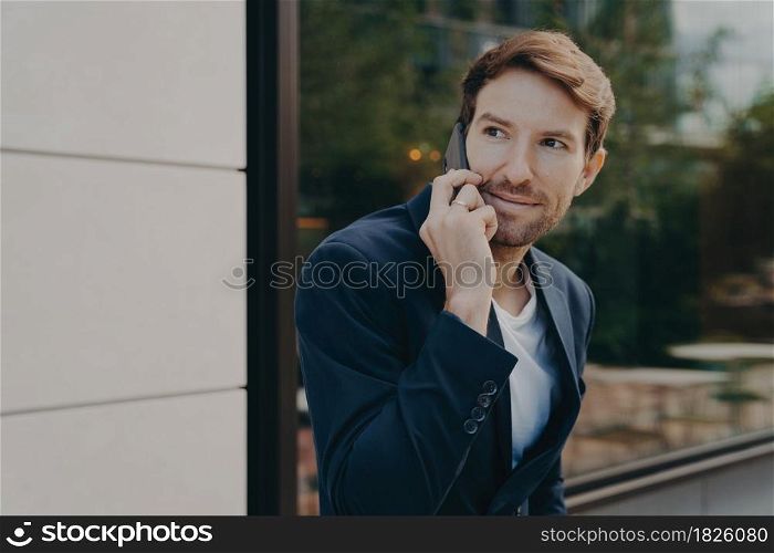 Business communication. Young handsome urban professional businessman listening to customer on cell phone, discussing something on smartphone, standing alone next to building with reflective window. Handsome urban professional businessman listening to customer on cell phone while standing outside