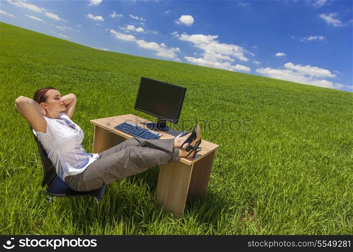 Business concept beautiful woman sitting relaxing day dreaming at desk feet up with computer in a green field with bright blue sky &amp; fluffy white clouds.