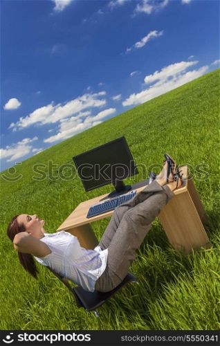 Business concept shot of a beautiful young woman businesswoman relaxing at an office desk &amp; computer in a green field with a bright blue sky &amp; white clouds.