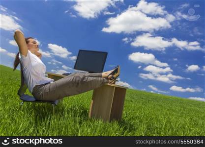 Business concept shot of a beautiful young woman businesswoman relaxing at an office desk &amp; computer in a green field with a bright blue sky &amp; white clouds.