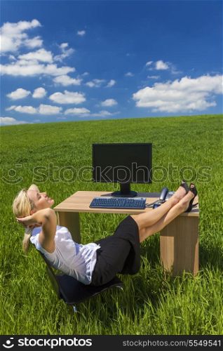 Business concept shot of a beautiful young woman businesswoman relaxing with her feet up at a desk with a computer in a green field with a bright blue sky