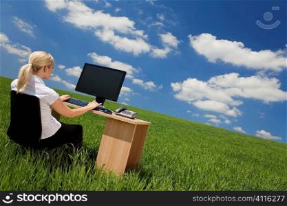 Business concept shot of a beautiful young woman sitting at a desk using a computer in a green field with a bright blue sky with fluffy white clouds. Shot on location.