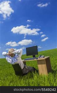 Business concept shot showing an older male executive relaxing at a desk with a computer in a green field &amp; blue sky complete with fluffy white clouds.