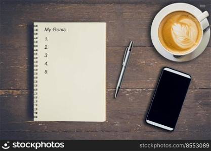 Business concept - Top view notebook writing My Goals, pen, coffee cup, and phone on wood table.