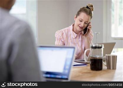 Business Couple Working From Home Sitting At Table During Pandemic Lockdown Using Mobile Phone