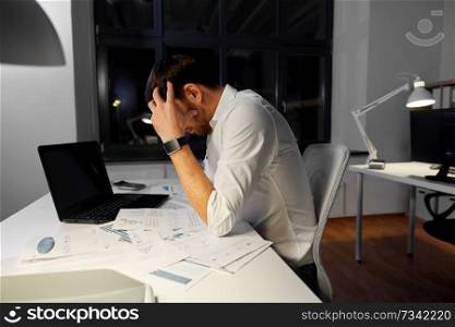 business, deadline and stress concept - stressed businessman with papers and laptop computer working at night office. businessman with papers working at night office