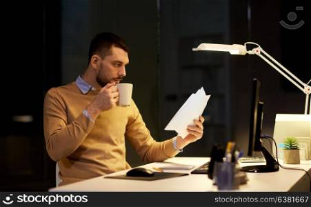 business, deadline and technology concept - man with papers working and drinking coffee at night office. man with papers and coffee working at night office