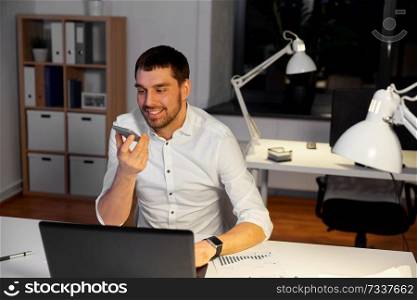 business, deadline and technology concept - smiling businessman with laptop computer using voice command recorder on smartphone at night office. businessman recording voice message on smartphone