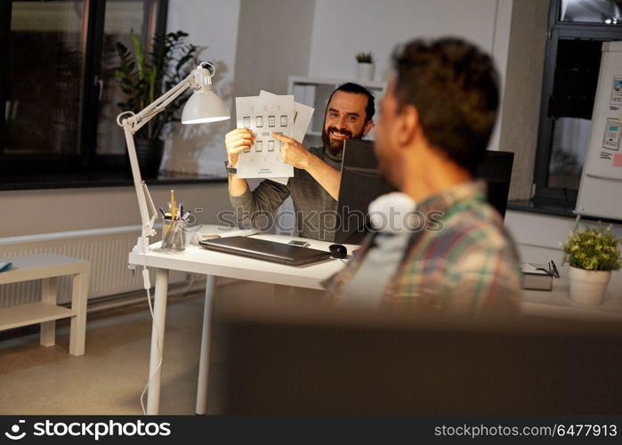 business, deadline and web design people concept - happy creative man showing papers to colleague at night office. creative man showing papers to colleague at office. creative man showing papers to colleague at office