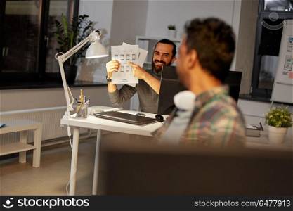 business, deadline and web design people concept - happy creative man showing papers to colleague at night office. creative man showing papers to colleague at office. creative man showing papers to colleague at office