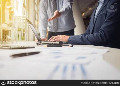 business documents on office table with laptop and digital tablet and stylus and two colleagues discussing data in the background.