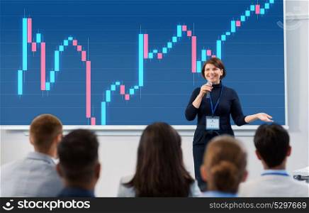 business, economy and people concept - smiling businesswoman or financier with forex chart on projection screen microphone talking to group of students at conference presentation or lecture. group of people at business conference or lecture