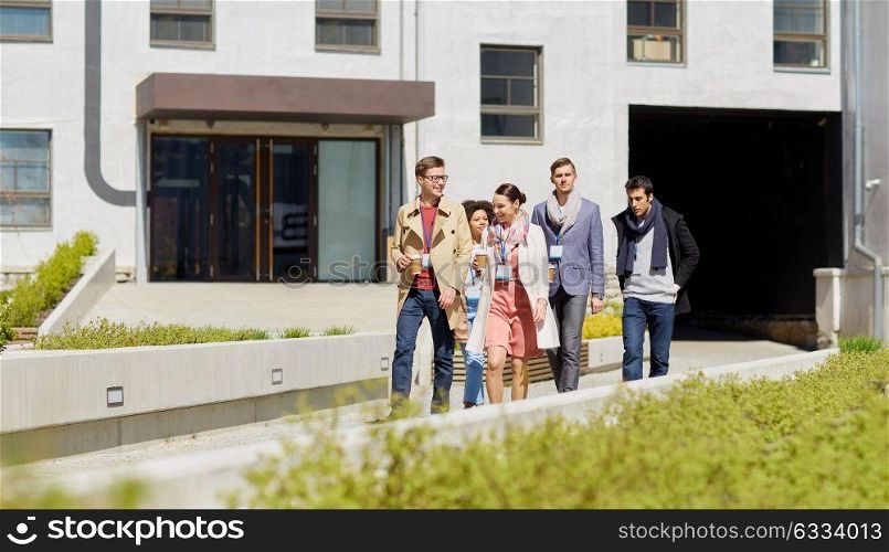 business, education and corporate concept - happy international group of office workers with conference badges drinking coffee and walking outdoors. office workers with coffee walking in city