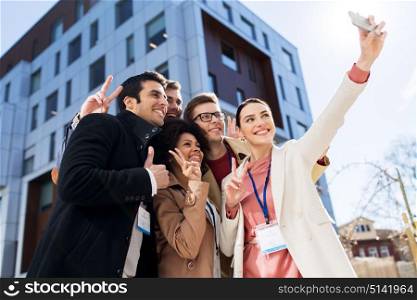 business, education and corporate concept - international group of people with conference badges taking selfie by smartphone on city street. happy people with conference badges taking selfie