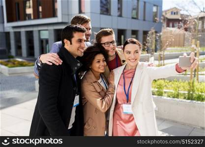 business, education and corporate concept - international group of people with conference badges taking selfie by smartphone on city street. happy people with conference badges taking selfie