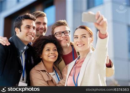 business, education and corporate concept - international group of people with conference badges and smartphone taking selfie on city street. business team with conference badges taking selfie