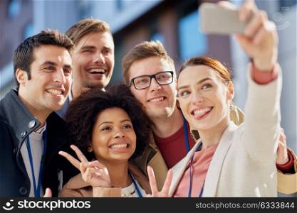 business, education and corporate concept - international group of people with smartphone taking selfie on city street and showing peace hand sign. business team with conference badges taking selfie