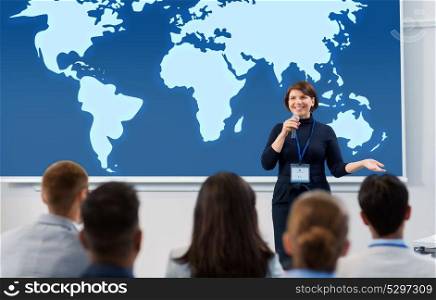business, education and people concept - smiling businesswoman or lecturer with microphone and world map on projection screen talking to group of students at conference presentation or lecture. group of people at business conference or lecture