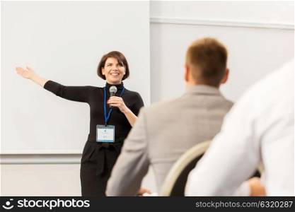 business, education and people concept - smiling businesswoman or lecturer with microphone talking to group of students at conference presentation or lecture and showing something on screen. group of people at business conference or lecture