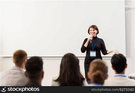 business, education and people concept - smiling businesswoman or teacher with microphone talking to group of students at conference presentation or lecture. group of people at business conference or lecture