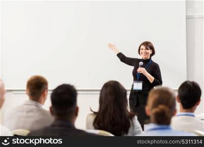business, education and people concept - smiling businesswoman or teacher with microphone talking to group of students at conference presentation or lecture and showing something on screen. group of people at business conference or lecture