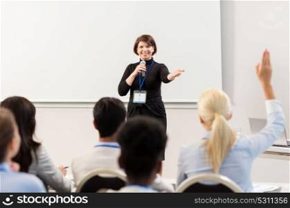 business, education and people concept - smiling businesswoman or teacher with microphone answering questions at conference presentation or lecture. group of people at business conference or lecture