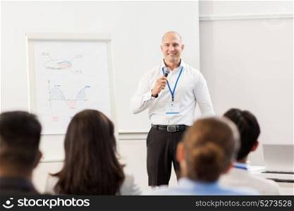 business, education and strategy concept - smiling businessman with microphone and charts on whiteboard talking to group of people at conference presentation. group of people at business conference