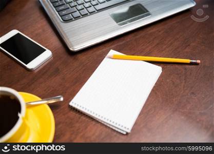 business, education and technology concept - close up of notebook with pencil, laptop, coffee and smartphone on table