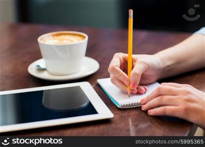 business, education, people, technology and lifestyle concept - close up of woman hands writing to notebook by pencil and tablet pc computer with coffee