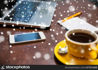 business, education, technology and object concept - close up of smartphone, coffee cup and laptop on table over snow