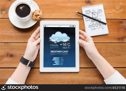 business, education, technology and people concept - close up of woman with weather forecast application on tablet pc computer screen, notebook and coffee on wooden table