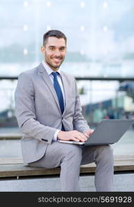 business, education, technology and people concept - smiling businessman working with laptop computer on city street