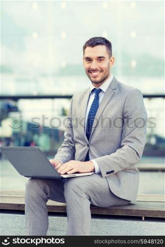 business, education, technology and people concept - smiling businessman working with laptop computer on city street