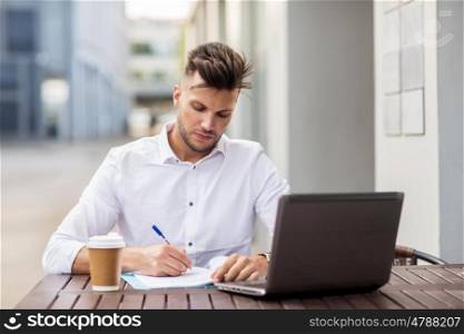 business, education, technology and people concept - young man with laptop computer, documents and coffee cup at city street cafe