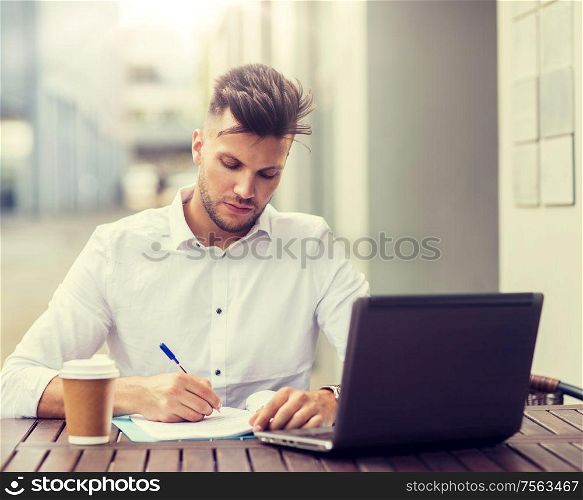 business, education, technology and people concept - young man with laptop computer, documents and coffee cup at city street cafe. man with laptop and coffee at city cafe