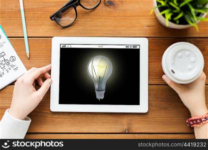 business, education, technology, people and inspiration concept - close up of woman with light bulb icon on tablet pc computer screen and coffee on wooden table