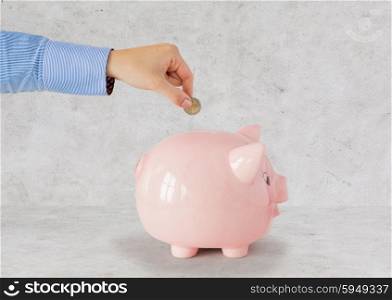 business, finance, investment, money saving and budget concept - close up of hand putting coin into piggy bank over gray concrete background