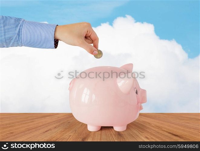 business, finance, investment, money saving and budget concept - close up of hand putting coin into piggy bank over blue sky and wooden floor background