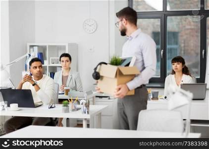 business, firing and job loss concept - fired male office worker with box of personal stuff looking at his sad colleagues. fired male office worker with personal stuff