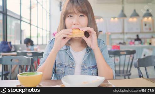 Business freelance Asian woman eating bread and drinking warm cup of coffee while sitting on table in cafe. Lifestyle smart beautiful female relax in coffee shop concepts.