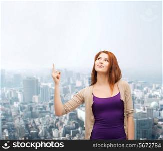 business, gesture and people concept - smiling teenage girl in casual clothes pointing finger up over city background