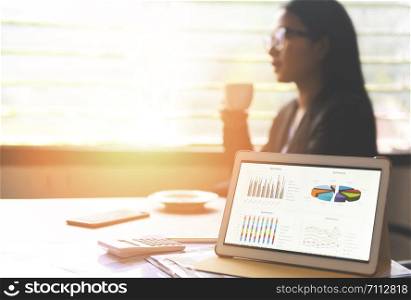 Business graphs chart on a tablet computer technology on the table and woman working in office with coffee cup / Sale report money analyzing