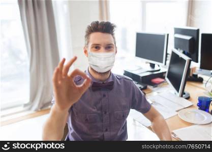 business, health and people concept - businessman or creative male office worker with computers wearing face protective medical mask for protection from virus disease and showing ok hand sign. male office worker in mask showing ok sign