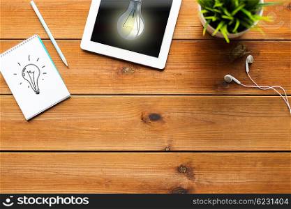 business, idea and technology concept - close up of tablet pc computer with light bulb on screen, notebook with scheme, pencil and earphones on wooden table