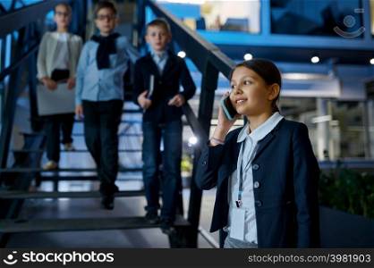 Business kids at office building hall. Little girl talking phone while partners standing on stairs. Business children at hall of office building