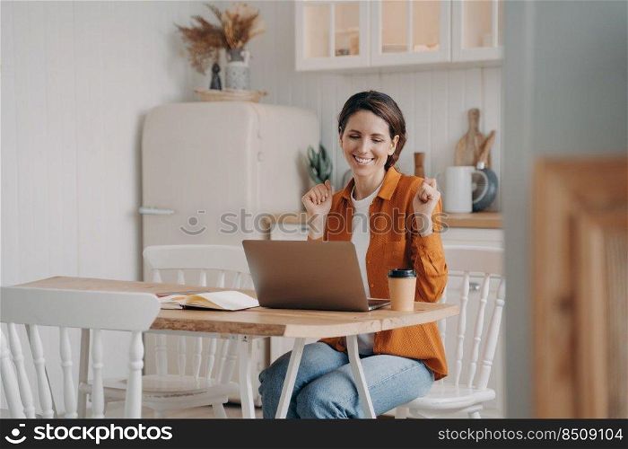 Business lady has online conference. Confident young woman wins. Girl is working in front of computer. European woman is happy. Distance work at the kitchen on quarantine concept.. Business lady has online conference. Confident woman wins. Distance work at kitchen on quarantine.