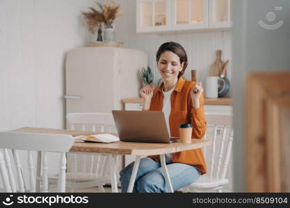 Business lady has online conference. Confident young woman wins. Girl is working in front of computer. European woman is happy. Distance work at the kitchen on quarantine concept.. Business lady has online conference. Confident woman wins. Distance work at kitchen on quarantine.