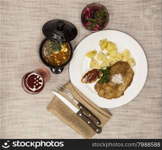 Business lunch : Petite Wiener schnitzel with boiled potatoes and ketchup. Served on a white porcelain plate with fork and knife on a wooden background. Beet salad and tomato, red pepper soup, sauce with olive oil, rosemary and smoked paprika with fork and a glass of juice and knife on a wooden background.